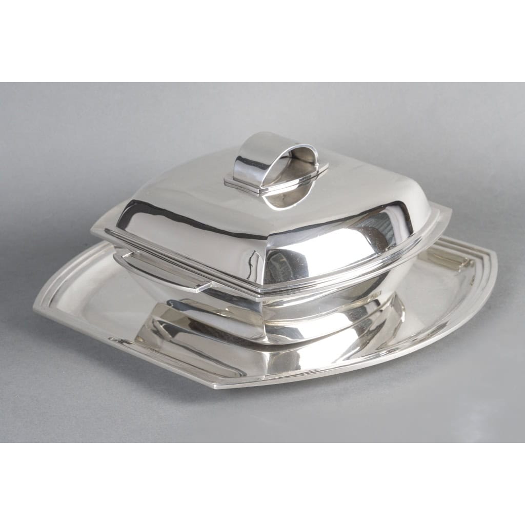 CHRISTOFLE - SOUPIERE MODERNISTE SUR SON PLATEAU EN ARGENT MASSIF ART DECO CHRISTOFLE - SOUPIERE MODERNISTE SUR SON PLATEAU EN ARGENT MASSIF ART DECO Centre de table formé d'une soupière couverte sur son plateau, de forme rectangulaire à fond plat et flanqué de deux anses latérales, le couvercle à doucine est surmonté d'une prise en demi-cercle. La soupière repose sur un plateau en argent aux mêmes formes incurvées. Dimensions : Soupière : 26 x 22 x 12 cm - base 16 x 16 cm plateau : 30 x 30,5 cm hauteur 1,5 cm Matière : Argent 1er titre 950/°°° Poids : 2 060 grammes Poinçon : MINERVE Epoque : ART DECO vers 1930 Orfèvres : CHRISTOFLE & RAVINET D'ENFERT pour le plateau Origine : France/Paris